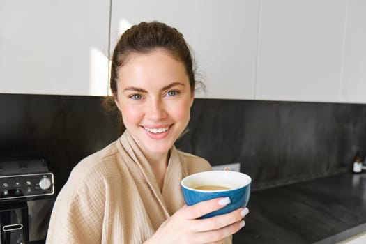 Good-looking young woman with cup of coffee, posing in the kitchen, smiling at camera, enjoys her morning at home, wearing cosy bathrobe.