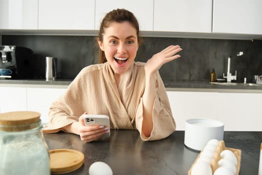 Attractive young cheerful girl baking at the kitchen, making dough, holding recipe book, having ideas.