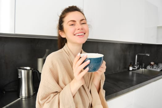 Beautiful young woman enjoys her morning mug of coffee, drinking tea from cup in the kitchen, wearing bathrobe and smiling at camera.