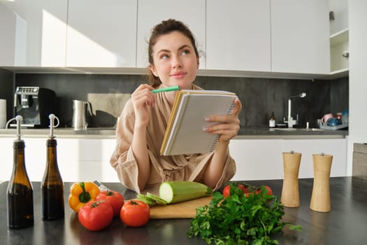 Portrait of woman checking recipe notes in notebook, standing in kitchen with vegetables, cooking food, preparing delicious salad from tomatoes, parsley and olive oil.