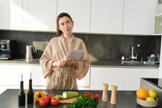 Portrait of creative young woman, cooking, holding recipe book, thinking while making meal, preparing food in the kitchen, standing in bathrobe.