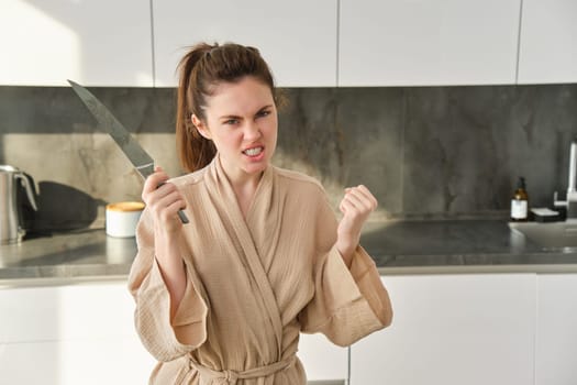 Portrait of annoyed woman with knife, angry while cooking in the kitchen, frustrated while doing house chores and preparing food for family, standing in bathrobe.