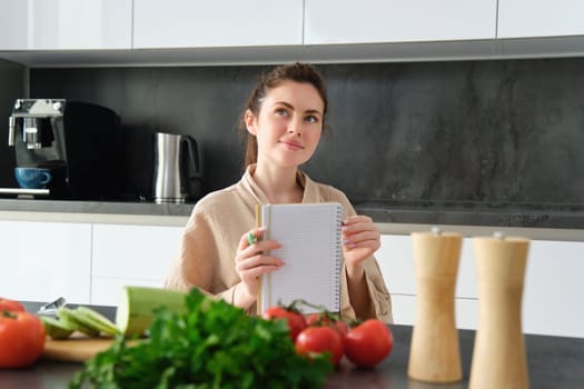 Portrait of happy young woman writes down menu for dinner, sits in the kitchen near vegetables, makes grocery list for shopping, poses in the bathrobe.