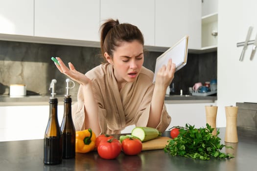 Portrait of woman cant cook, looking confused while making meal, holding recipe book, checking grocery list and staring frustrated at camera, standing near vegetables in the kitchen.