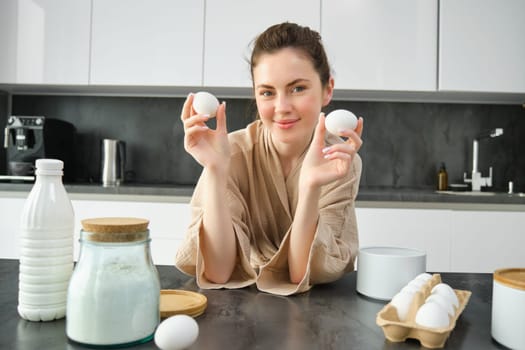 Attractive young cheerful girl baking at the kitchen, making dough, holding recipe book, having ideas.