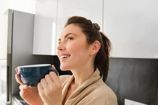 Close up portrait of dreamy, smiling young woman, drinks coffee and looks thoughtful, stands in kitchen.