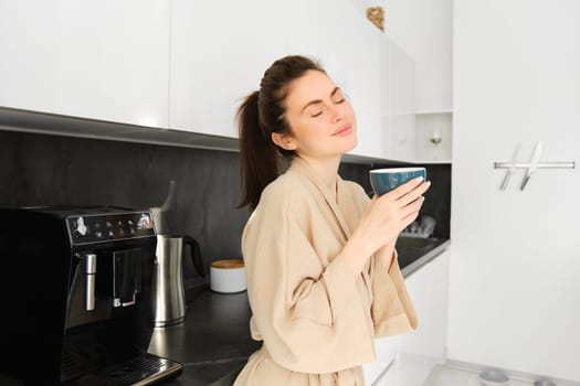Portrait of woman standing near coffee machine, preparing morning cup of cappuccino, standing in kitchen, wearing bathrobe.