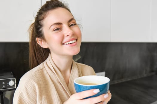 Portrait of smiling woman drinks coffee, stands in the kitchen and enjoys delicious cup of cappuccino in the morning, looks happy at camera.