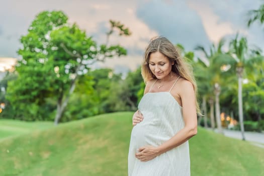 Pregnant woman hugging her tummy standing outdoors surrounded by nature. Pregnancy, expectation, motherhood concept.