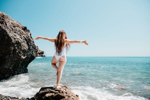 Woman sea yoga. Back view of free calm happy satisfied woman with long hair standing on top rock with yoga position against of sky by the sea. Healthy lifestyle outdoors in nature, fitness concept