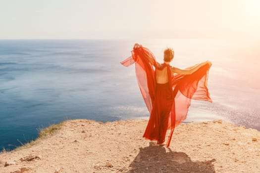 Side view a Young beautiful sensual woman in a red long dress posing on a rock high above the sea during sunrise. Girl on the nature on blue sky background. Fashion photo.