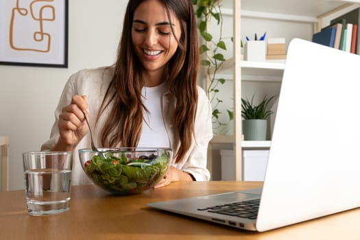 Caucasian woman eating healthy salad for lunch while working with laptop at home office. Healthy lifestyle concept.