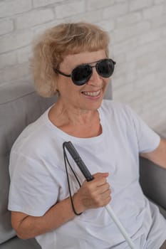 An elderly blind woman wearing sunglasses and with a tactile cane sits on the sofa