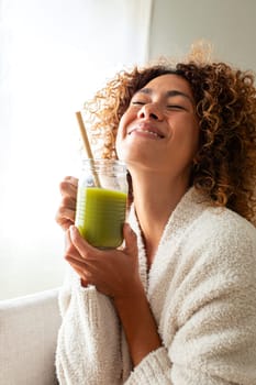 Happy multiracial woman enjoying healthy green juice at home. Female doing detox drinking green smoothie. Vertical image. Healthy lifestyle concept.