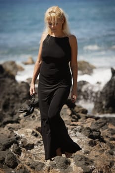 Beautiful blonde woman wandering along rocky seashore in black dress while holding dress shoes