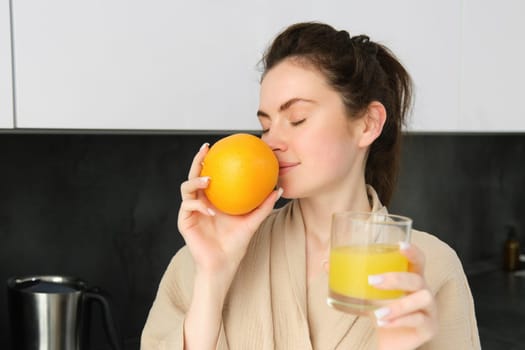 Close up portrait of young brunette, smells fresh orange, drinking juice from glass, enjoying healthy start of morning, wearing bathrobe.