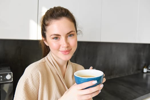 Good-looking young woman with cup of coffee, posing in the kitchen, smiling at camera, enjoys her morning at home, wearing cosy bathrobe.