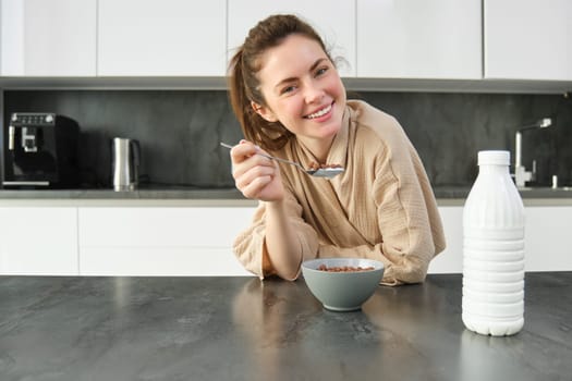 Laughing, beautiful woman in bathrobe, eats cereals with milk, holding spoon, smiling and looking happy, posing in kitchen and leaning on worktop.