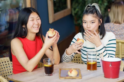 Cheerful young Asian female friends in casual clothing gathering at wooden table with drinks and eating yummy food while having lunch together in cafe