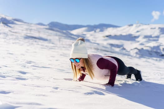 Cheerful young female in warm clothes and sunglasses lying in Chaturanga pose on white snowy slope of mountain on sunny winter day with blue sky