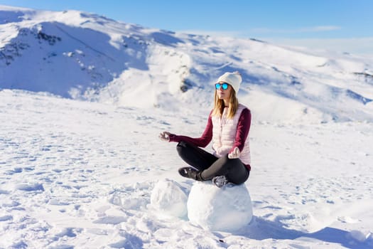 Full body of young female traveler in casual clothes and sunglasses sitting on snow in lotus pose and looking away on sunny winter day