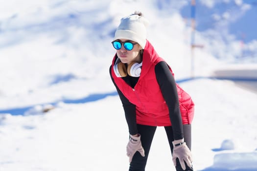 Young female in headphones and warm clothes leaning on knees while taking break from run against snowy mountains