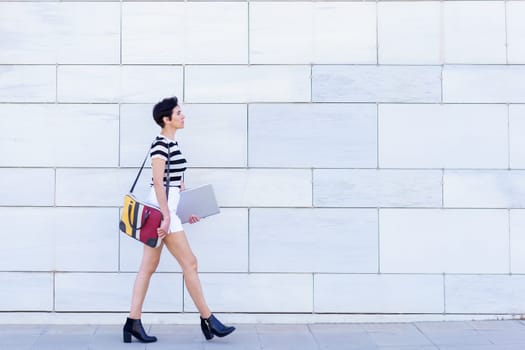 Full body side view of pensive young female in striped t shirt and shorts with bag and laptop walking on city street in daytime