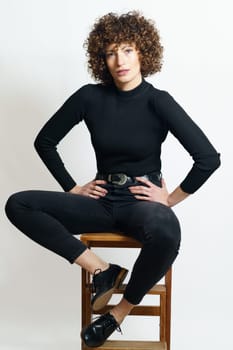 Full body of young confident female, model in black outfit relaxing on stool with steps while placing hands on his waist on seat and looking at camera against gray backdrop