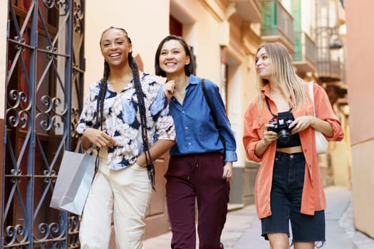 Cheerful young multiracial women in trendy clothes laughing while passing by building wall on blurred background