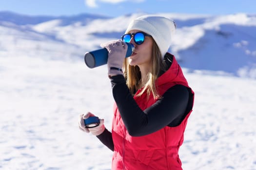 Side view of young female in warm clothes, beanie wool cap polarized sunglasses drinking warm water from thermos bottle and looking away while standing on snowy mountain slope in daylight
