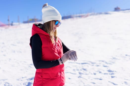 Side view of young female in warm clothes, beanie cap polarized sunglasses looking down at wristwatch while standing on snowy mountain slope in daylight against blurred landscape and blue sky