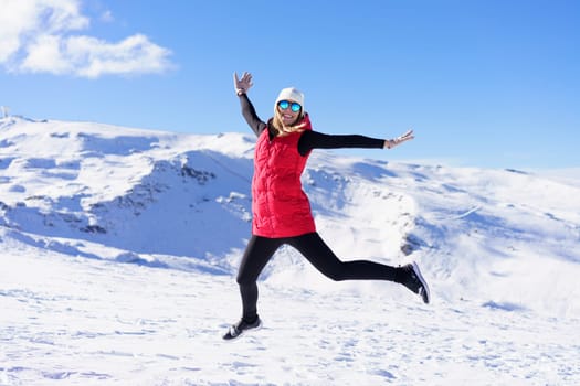 Full body of excited young female in vest cap, polarized sunglasses looking at camera while leaping up in air with stretched arms against snowy mountains and blue sky in daylight
