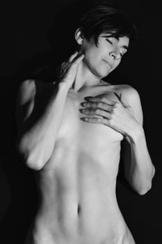 Black and white of sensual adult female with short dark hair covering naked breast and touching neck with closed eyes in studio