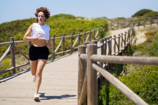 Full body of fit young sportswoman in activewear running along wooden walkway in countryside on sunny summer day