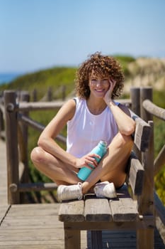 Young female athlete in sportswear smiling and looking at camera while sitting on wooden pier with bottle of water against blurred background