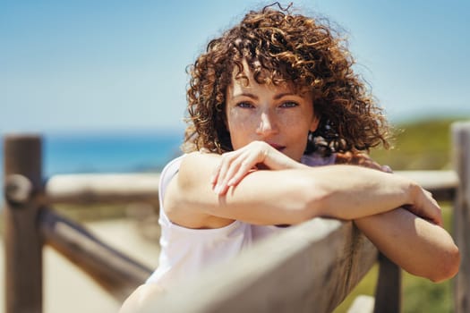 Calm young female with curly hair in white outfit leaning against fence and looking at camera while relaxing alone near blurred background of sea