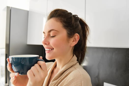 Portrait of happy young woman starts her morning with mug of coffee, drinking tea from cup, standing in the kitchen, smiling cheerfully.