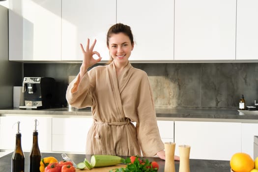Portrait of smiling brunette woman cooking healthy dinner, showing okay sign, wife standing in bathrobe in kitchen, preparing food, chopping vegetables on board.