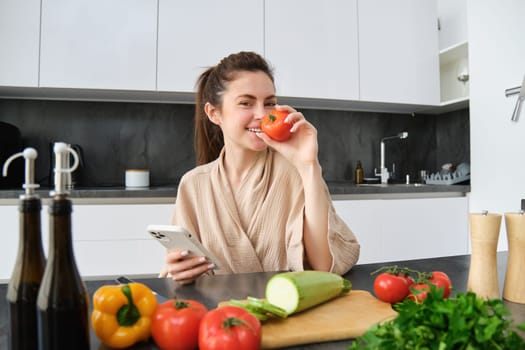 Portrait of young woman ordering groceries on smartphone app, holding tomato, sitting near chopping board with vegetables. Girl looking for recipe online, using mobile phone.