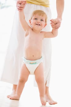 Mother helping baby walk indoors smiling