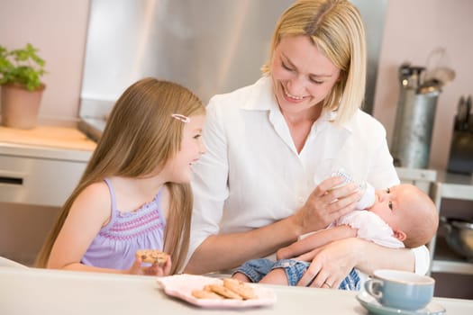 Mother feeding baby in kitchen with daughter eating cookies and smiling