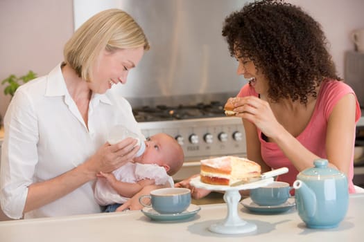 Mother and baby in kitchen with friend eating cake and smiling