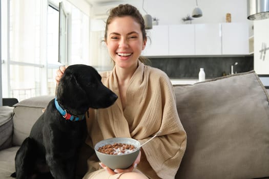Happy mornings. Beautiful woman enjoying her morning, sitting on sofa, eating breakfast and playing, cuddling with dog, puppy asks for small bite of cereals.