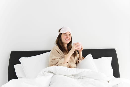 Portrait of beautiful woman enjoys her morning cup of coffee from bed, sitting in bedroom wearing sleeping mask and pyjamas, relaxing.