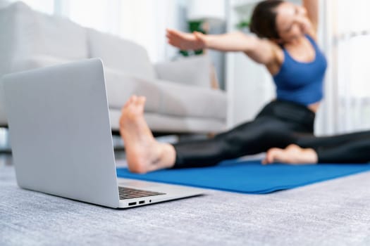 Focused laptop on the floor showing online exercise training video, while sporty athletic woman concentrate on warm-up and stretching routine in blurred background. Vigorous