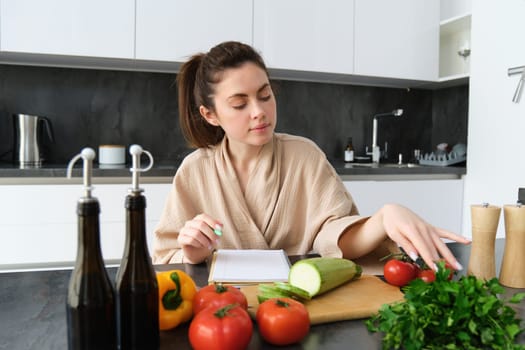 Portrait of woman writing down list of groceries, making notes in recipe, sitting in kitchen near vegetables, preparing dinner menu.