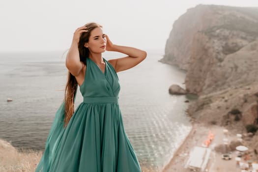 Side view a Young beautiful sensual woman in a mint long dress posing on a volcanic rock high above the sea during sunset. Girl on the nature on overcast sky background. Fashion photo