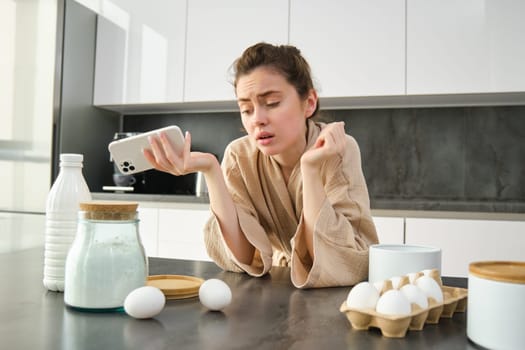 Attractive young cheerful girl baking at the kitchen, making dough, holding recipe book, having ideas.