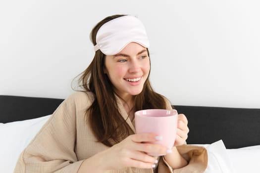 Happy young woman starting her morning with cup of coffee in bed, smiling and enjoying her lazy day, has sleeping mask on forehead.