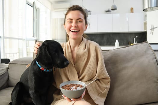 Home and lifestyle concept. Beautiful young woman with dog, sitting on sofa, eating cereals and hugging her puppy, girl having breakfast in bathrobe.